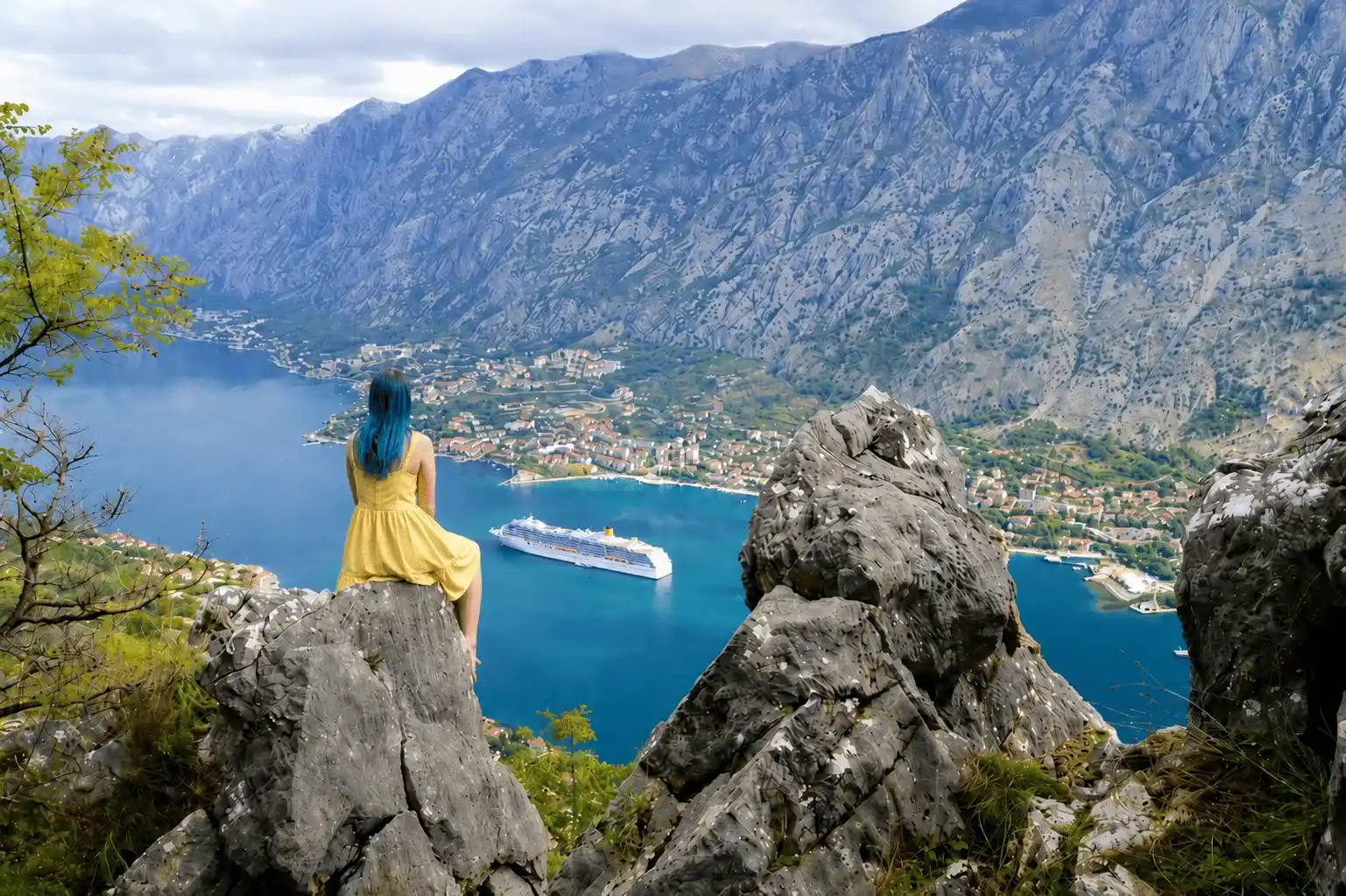 Žena sjedi na stijenama Vrmca i posmatra Kotor i kruzer u Bokokotorskom zalivu.