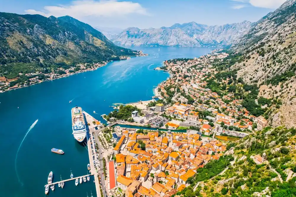 Panoramski pogled na Stari grad Kotor, luku sa kruzerom i Bokokotorski zaliv između planina.