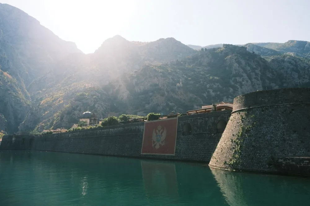 Gradske zidine Kotora uz more, sa crnogorskom zastavom i planinama u pozadini.